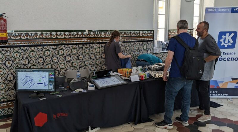 General view of the KDE booth at Akademy-es/OpenSOurthCode 2023. On the left, there is table loaded with screens, tablets, phones and consoles, on the right we have merch, such as t-shirts, mugs, caps, etc.